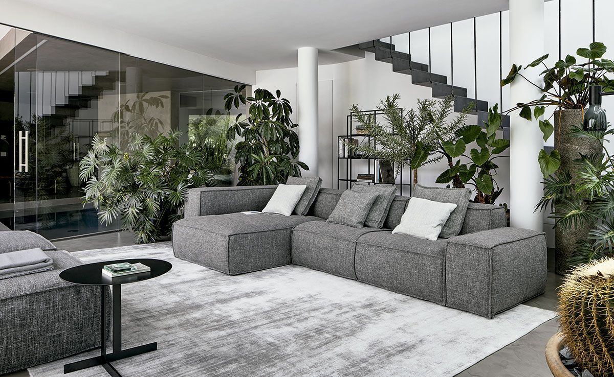 Sala de estar moderna con sofá seccional gris, escalera, plantas grandes y paredes de vidrio.
