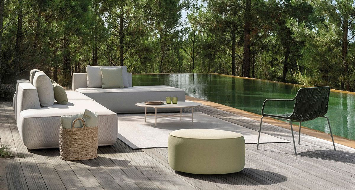 Patio exterior moderno con asientos blancos y beige, otomana redonda y vista al bosque.
