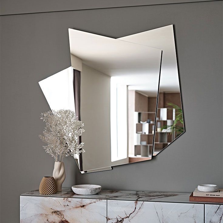 Espejo abstracto moderno sobre una consola de mármol con jarrón, cuenco y ramas decorativas en una habitación gris.