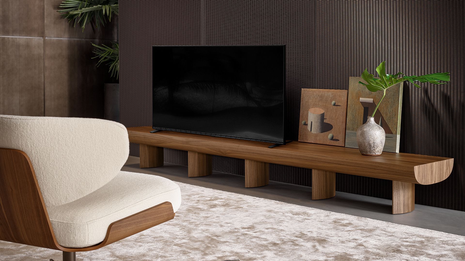 Sala de estar moderna con un televisor sobre una consola de madera, silla color crema, alfombra y plantas