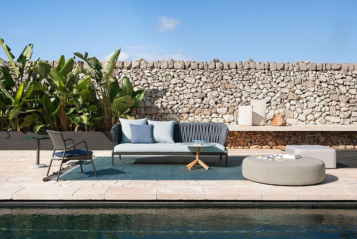 Patio moderno junto a la piscina con sofá gris, sillas, maceta y muro de piedra detrás de una piscina turquesa.