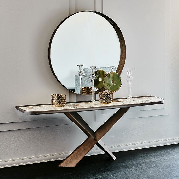 Mesa consola moderna con espejo redondo, decoración de cristal y vegetación sobre una pared blanca.