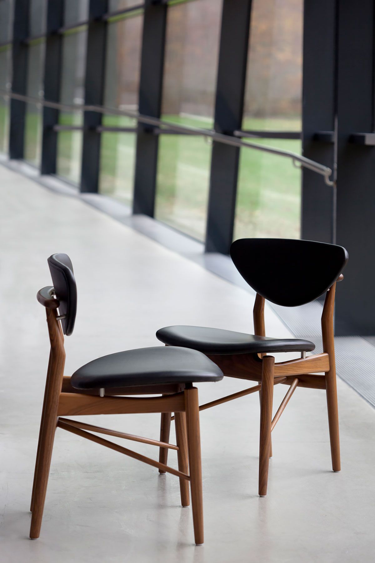 Dos sillas de madera con cojines negros en un pasillo moderno cerca de grandes ventanales.