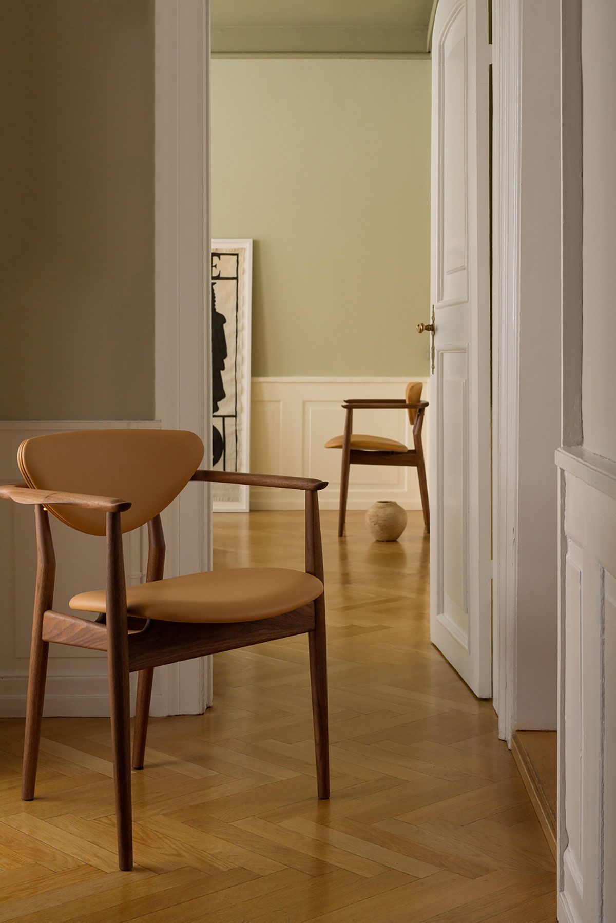 Pasillo con poca luz y sillas de madera que conduce a una habitación luminosa con una mesa y una silla.