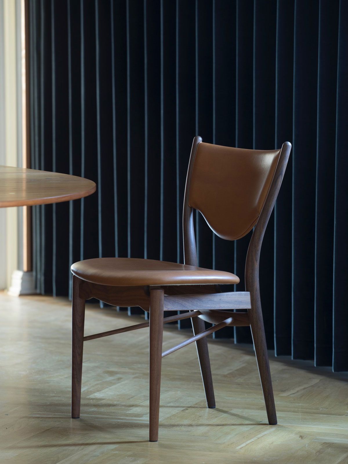 Silla de cuero marrón junto a una mesa redonda en una habitación con poca luz y cortinas oscuras.