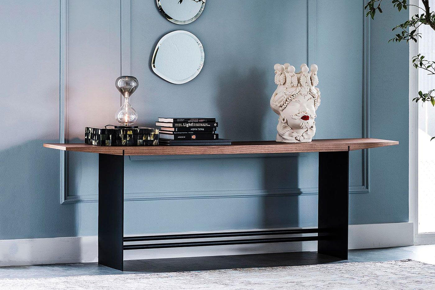 Consola de madera con elementos decorativos sobre una pared azul claro, con espejos, libros y una escultura blanca.