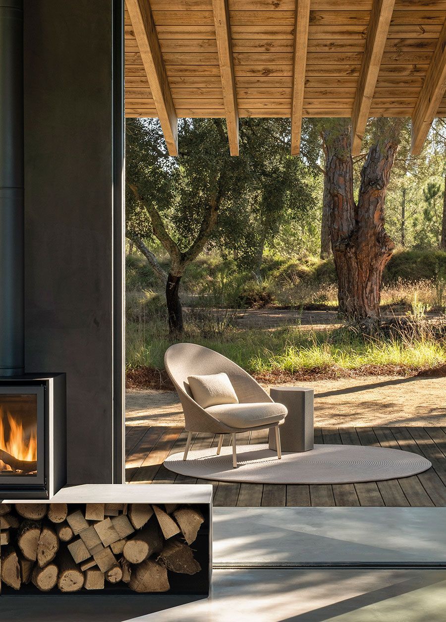 Patio moderno con una silla blanca, una alfombra redonda y una chimenea interior junto a una pila de leña.
