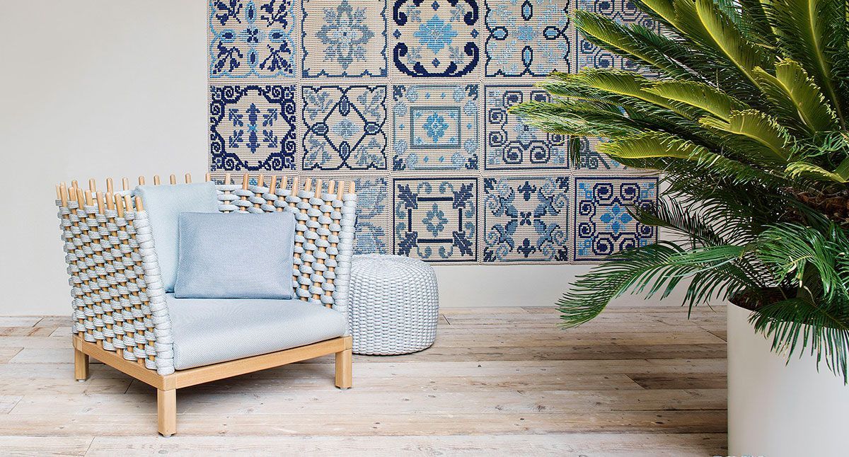 Silla de exterior con cojín azul junto a una pared de azulejos y una palmera en maceta en una terraza de madera soleada.