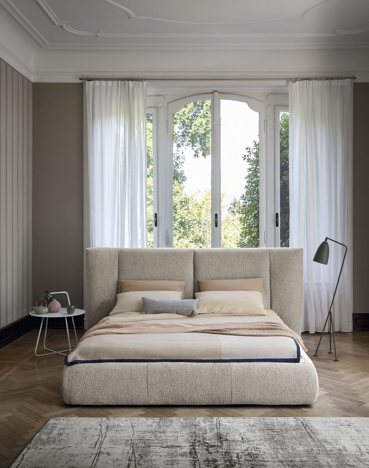Habitación luminosa con una cama grande, ventanas altas, cortinas transparentes y una lámpara de pie a la derecha.