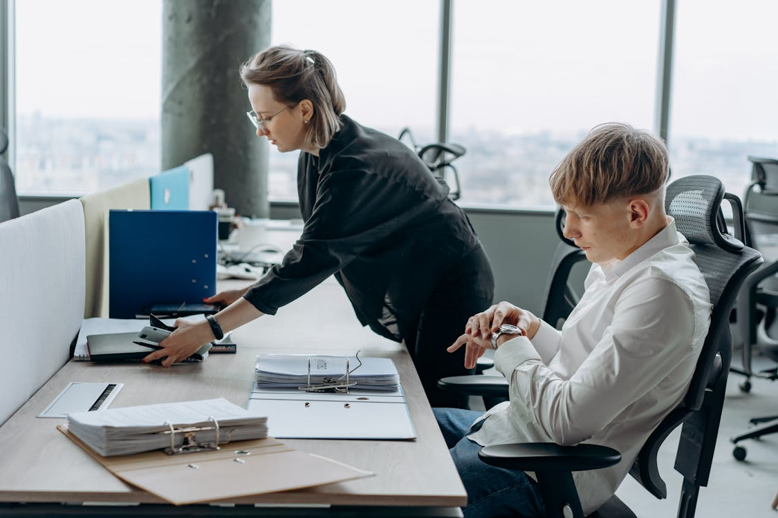 Manager assigning tasks while seated, employee checks time at desk, reflecting the need for employee engagement solutions.

