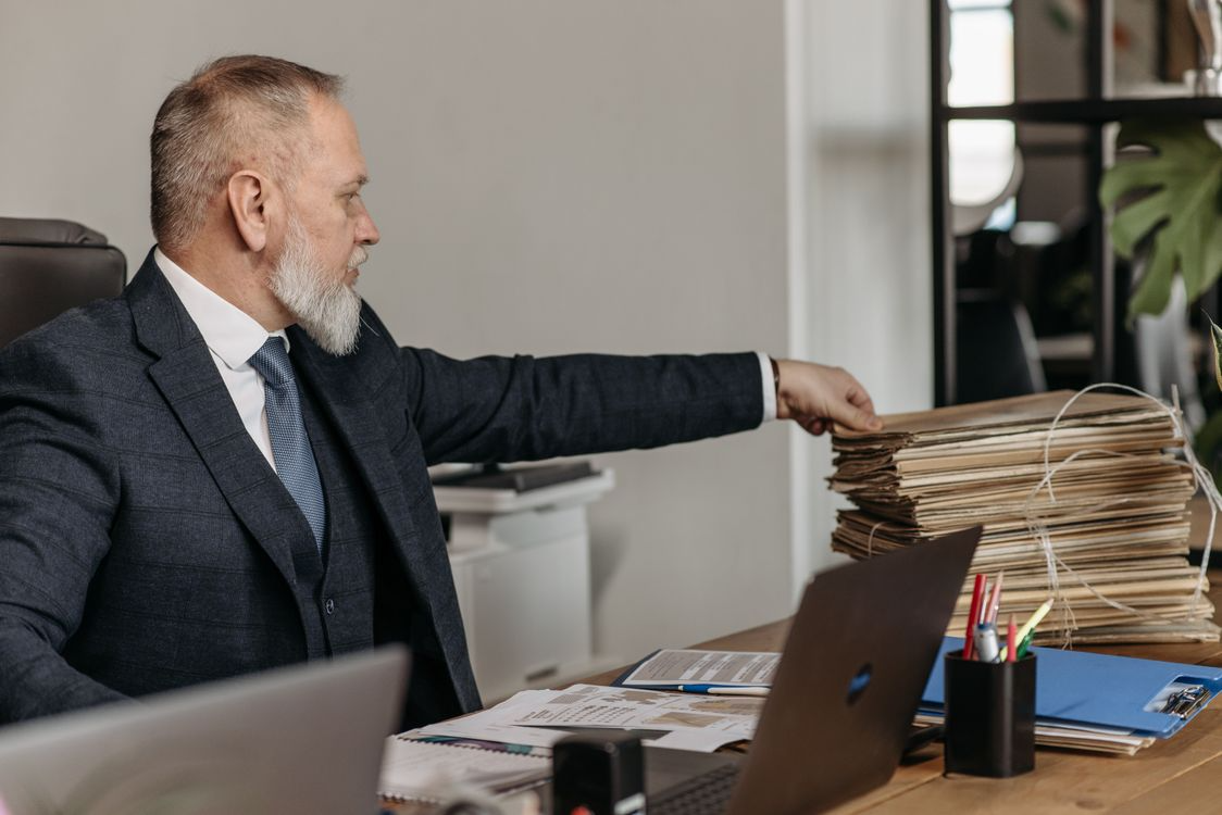 Senior business professional in a suit reaching toward a large stack of paper files on an office desk beside a laptop, illustrating heavy documentation workload and administrative processing.
