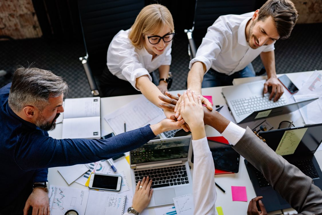 Team of professionals stacking their hands together in a collaborative huddle over a workspace with laptops and documents.

