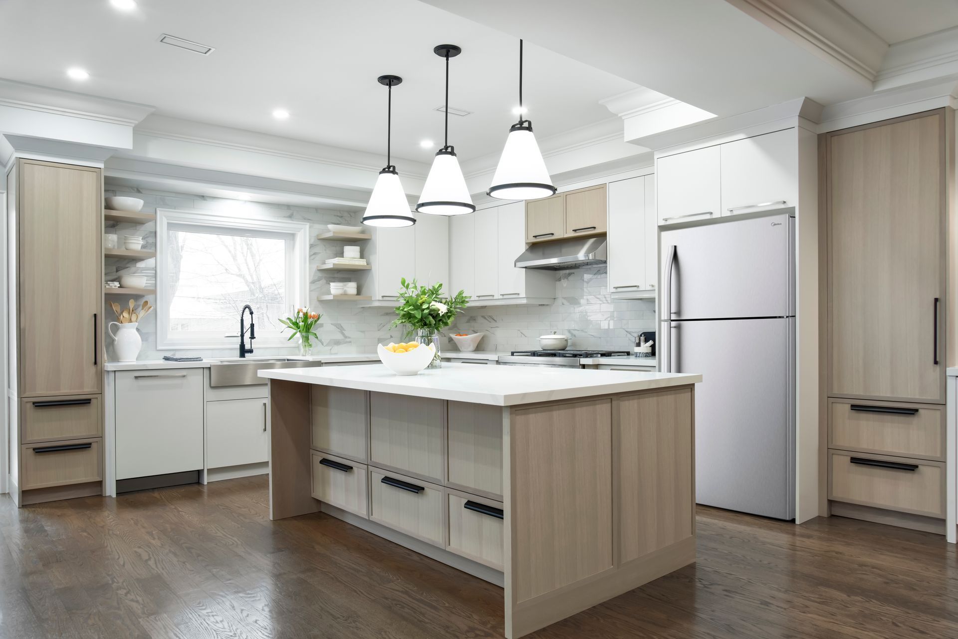 Modern kitchen with light wood cabinets, white island, stainless steel fridge, and pendant lights.
