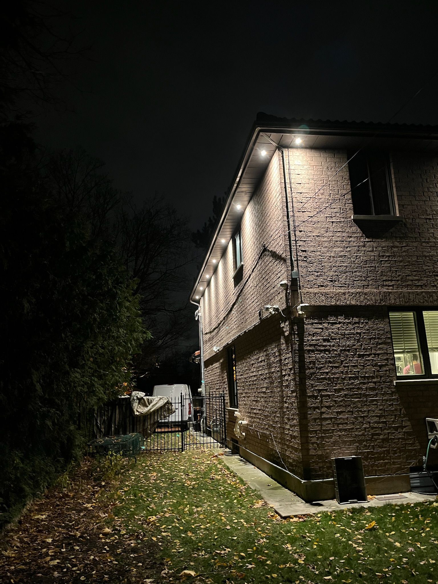 A two-story brick house at night, illuminated by soffit lights, with a dark lawn scattered with fallen leaves.