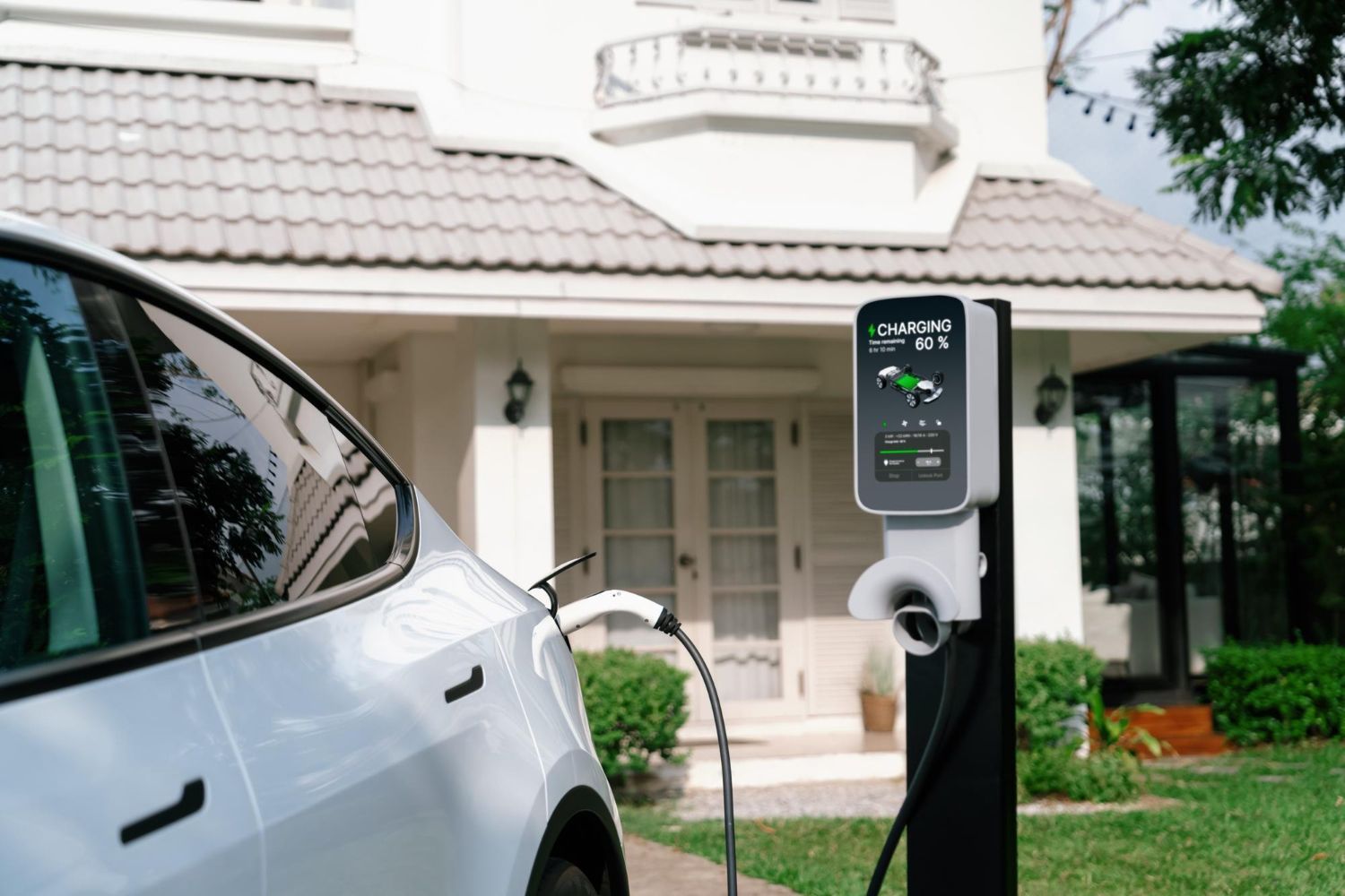 A white electric vehicle parked at a home residence is connected to a charging station displaying an active charge.