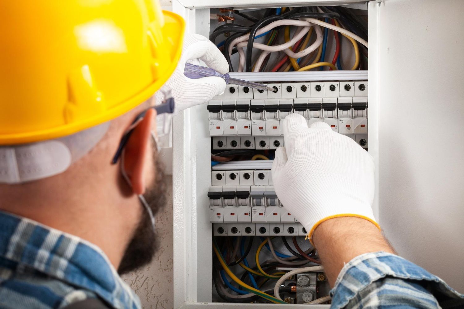 An electrician wearing a yellow hard hat and gloves inspects an open circuit breaker panel with a test tool.