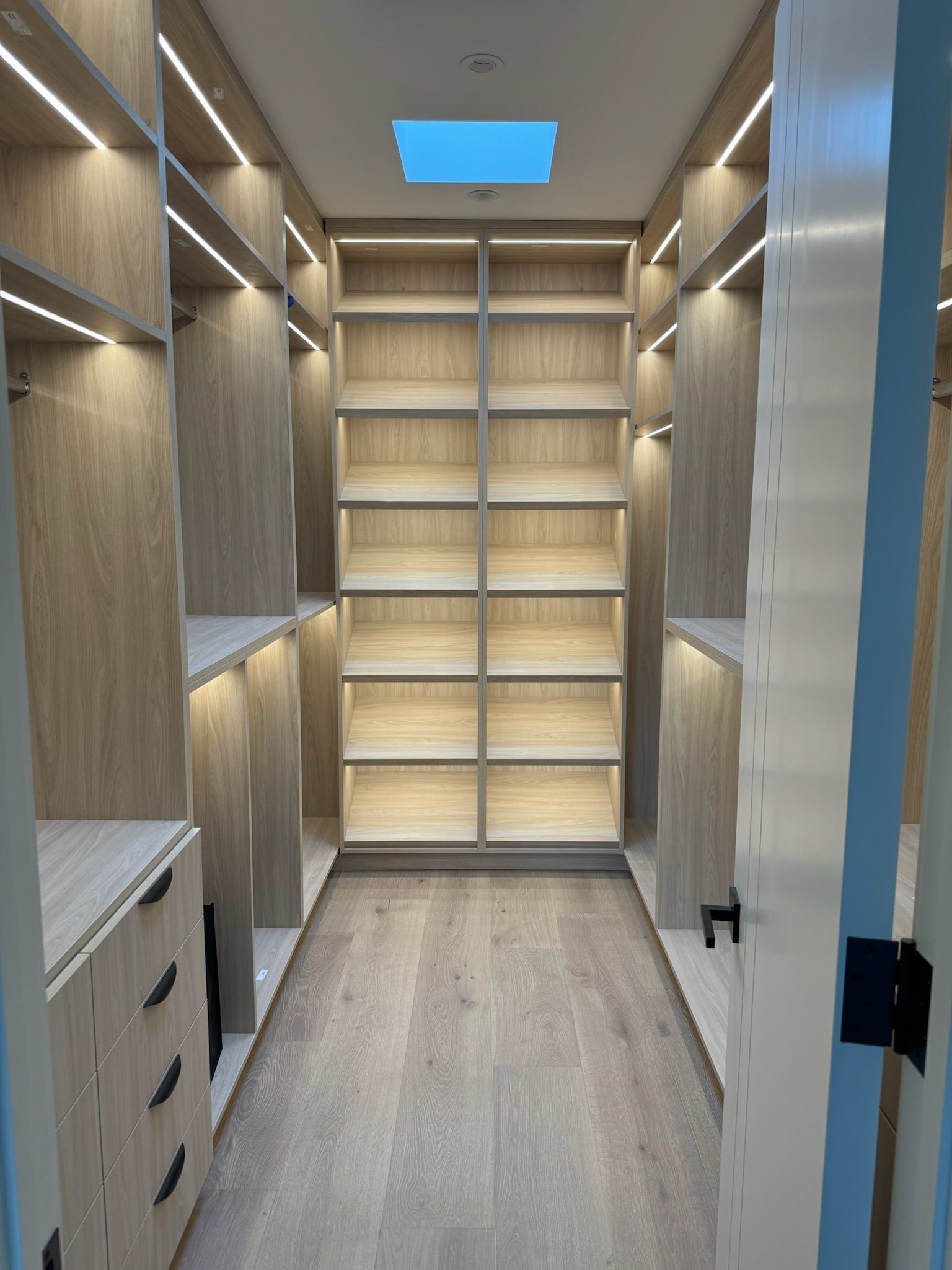 A modern, walk-in closet featuring light wood shelves, built-in drawers, vertical LED strip lighting, and a skylight.