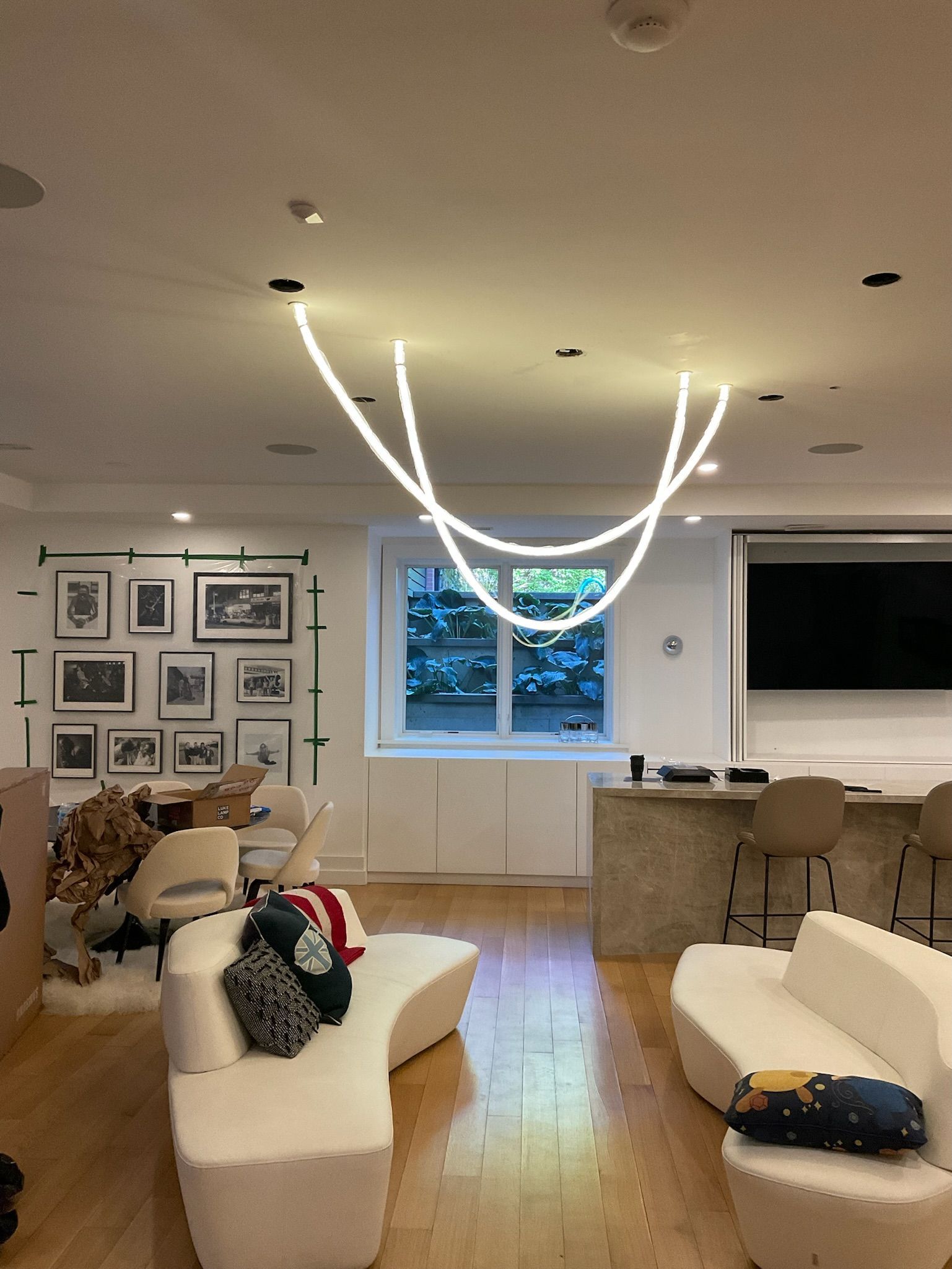 A modern, open-plan living room with curved white sofas, a concrete bar, light wood floors, and a looping pendant light.