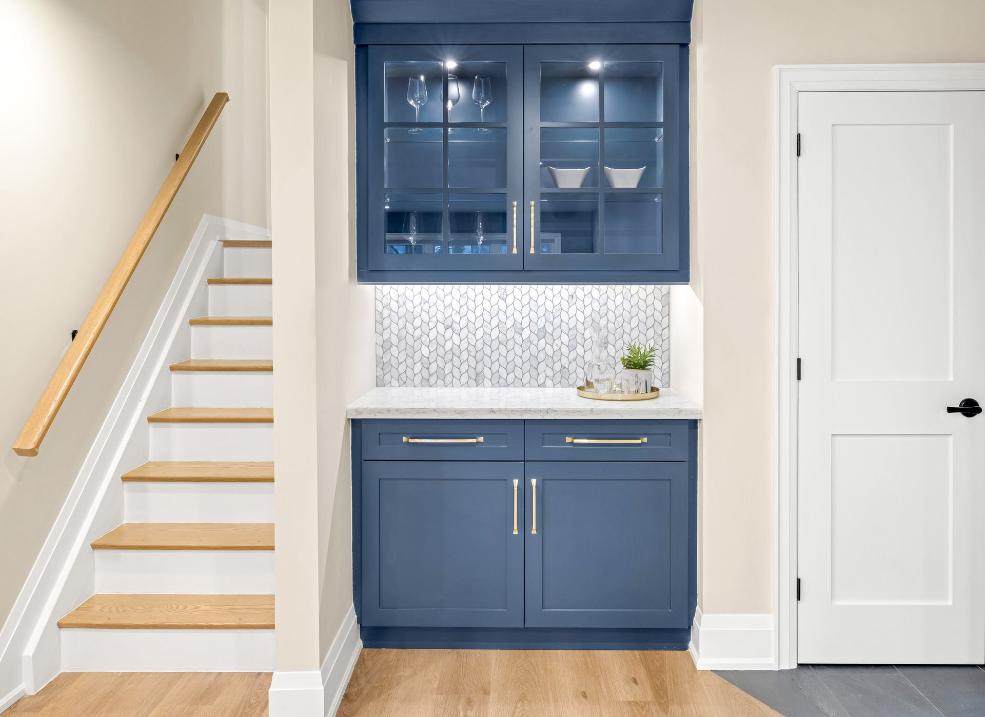 Blue built-in bar with upper glass cabinets, countertop, and lower cabinets next to stairs and a white door.
