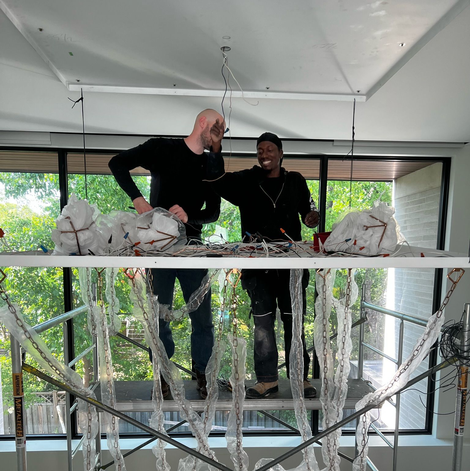 Two people working on a sculpture attached to scaffolding. Both are smiling. Large window in background.
