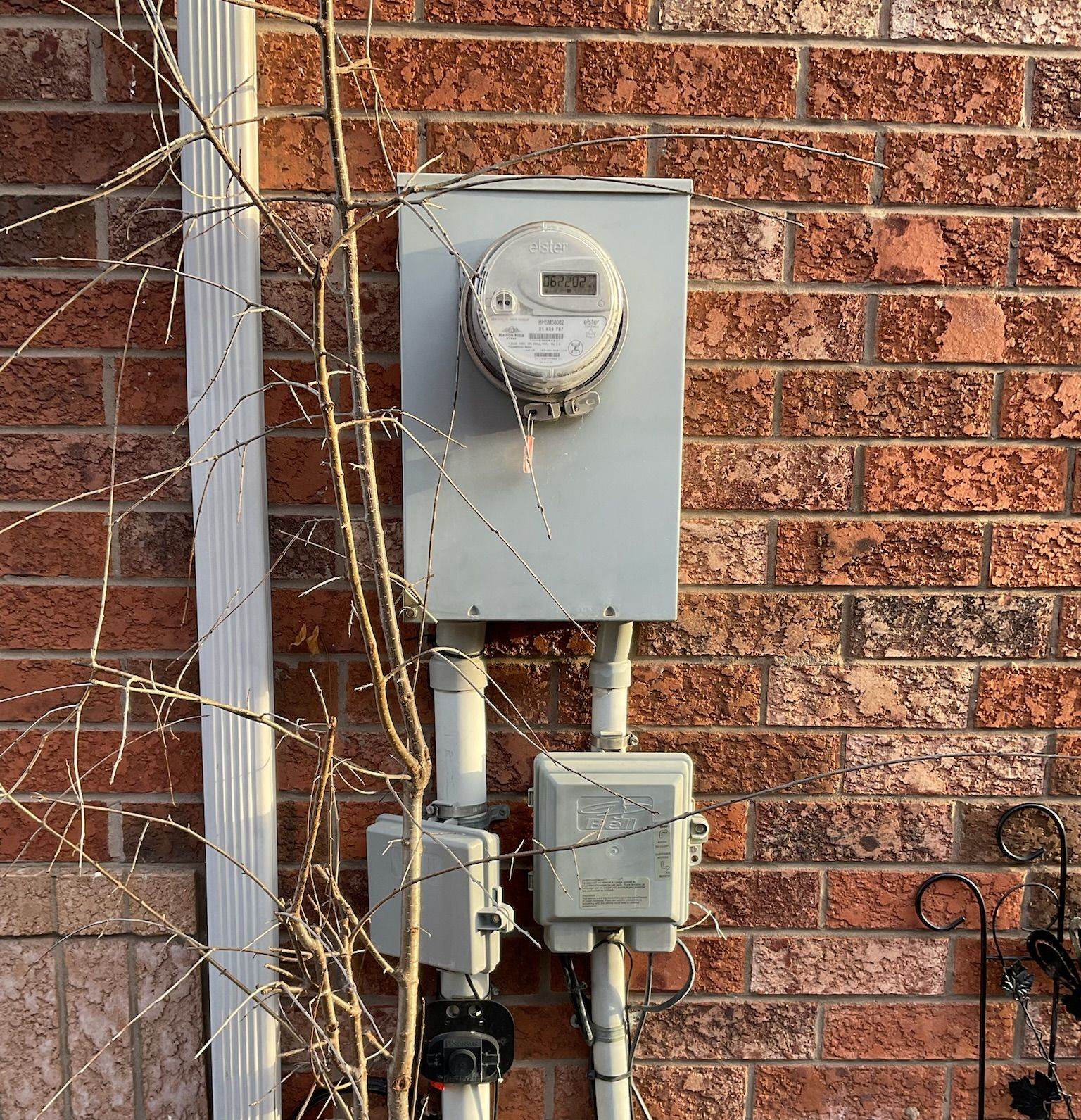 Gray electric meter and boxes on a brick wall with a gutter and a bare tree branch.