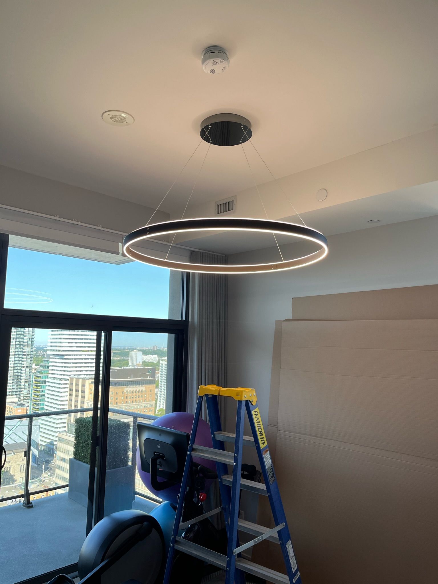 Circular LED chandelier hanging from a ceiling. A ladder leans against a wall, and a window with a city view is visible.