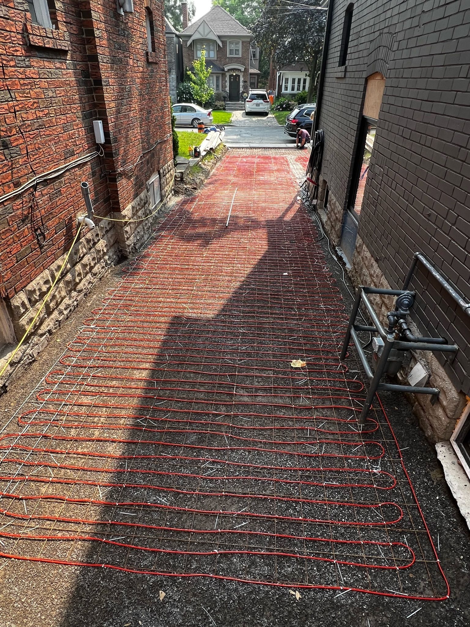 Brick-paved alleyway with red radiant heating tubes. Brick buildings line both sides, leading to a street with parked cars.