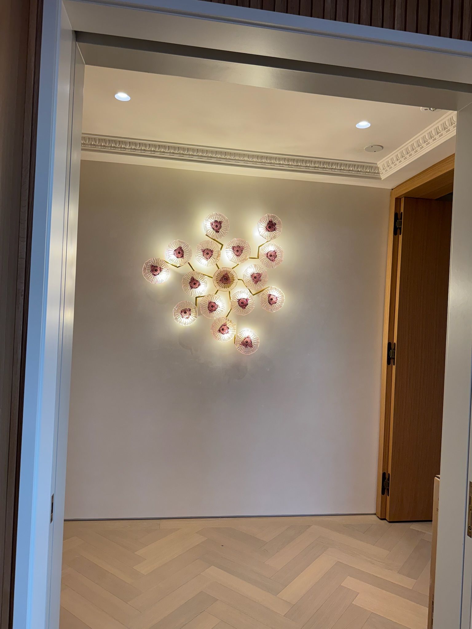 Hallway with floral-shaped wall sconce, light-colored walls, herringbone floor, and wooden doorframe.