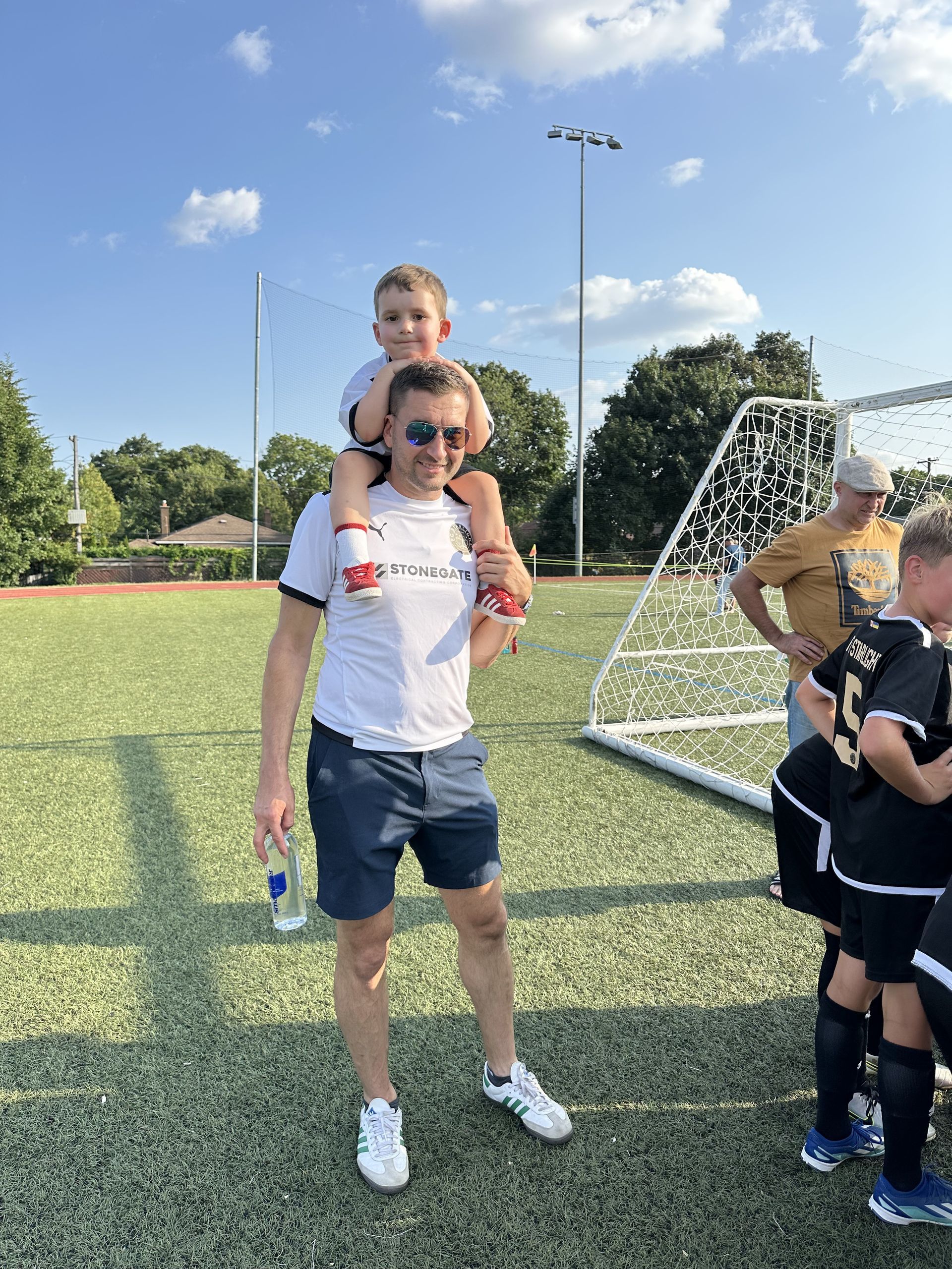 Man carries a child on his shoulders on a soccer field. They are smiling. Other people stand nearby.
