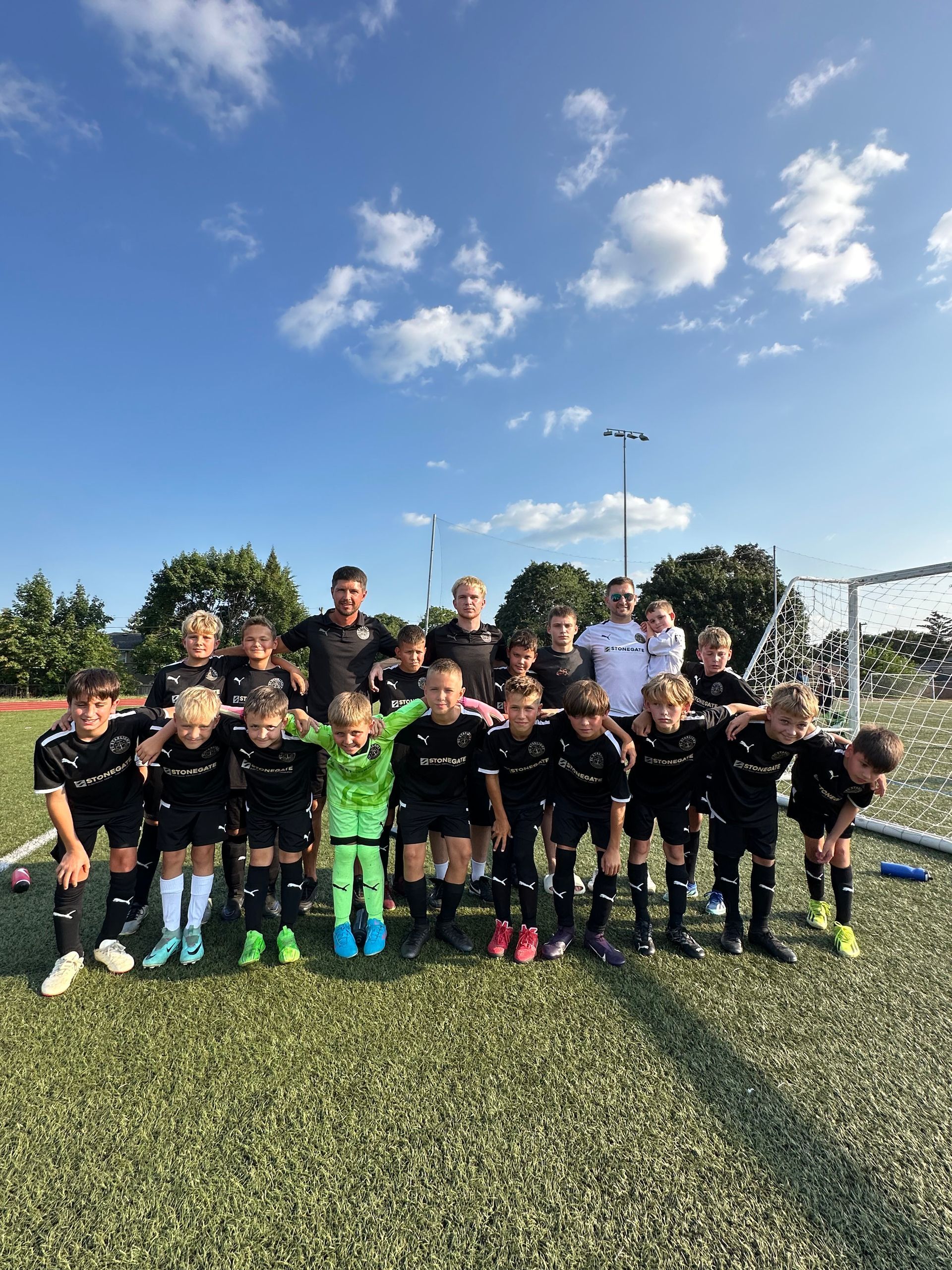 Soccer team in black uniforms and coaches pose on green field under a blue sky.