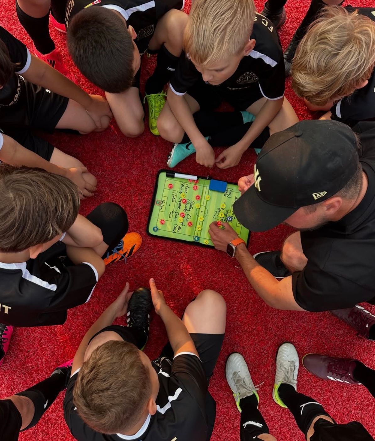 Soccer coach shows strategy on a clipboard to a team huddle on a red field. Players wear black uniforms.