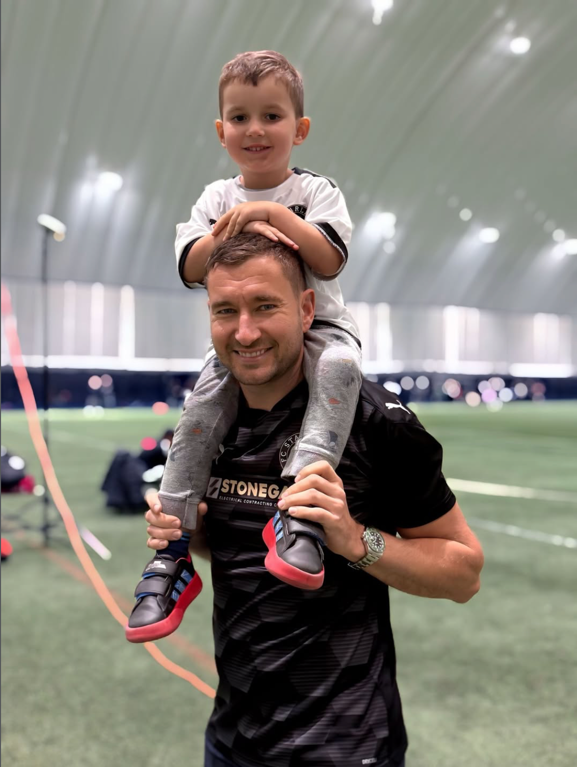 Man carrying a child on his shoulders at an indoor soccer field. The child wears a sports jersey. Both smiling.