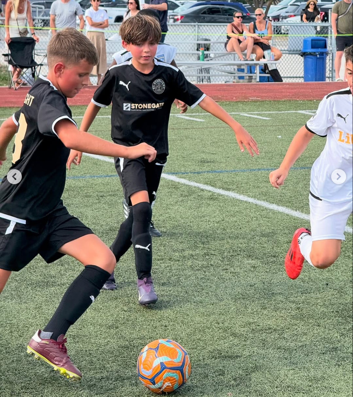Boys playing soccer on a green field. Two in black, one in white. A player dribbles the ball.