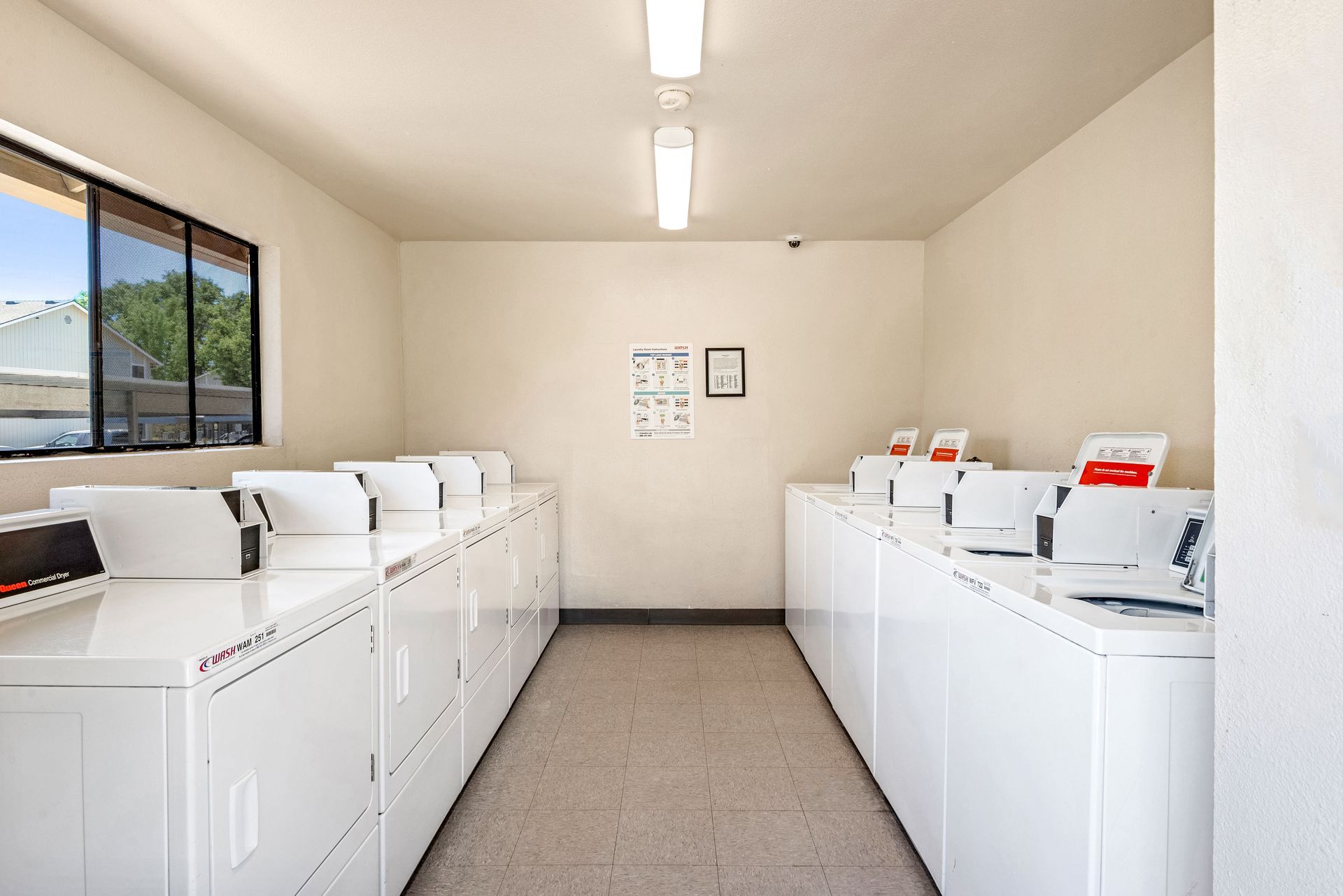 Laundry room with rows of white washers and dryers, beige walls, and a window.