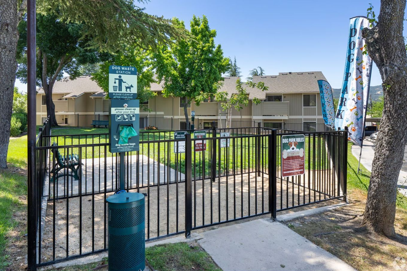Fenced dog park with signs, a trash can, and a bench; located in front of apartments with green trees.