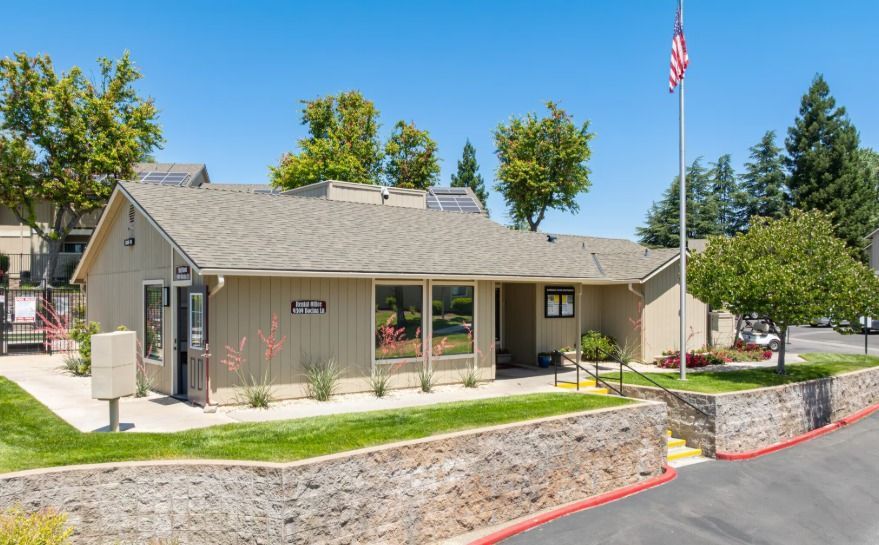 Tan building with a US flag, grassy area, and stone wall. Clear blue sky in the background.