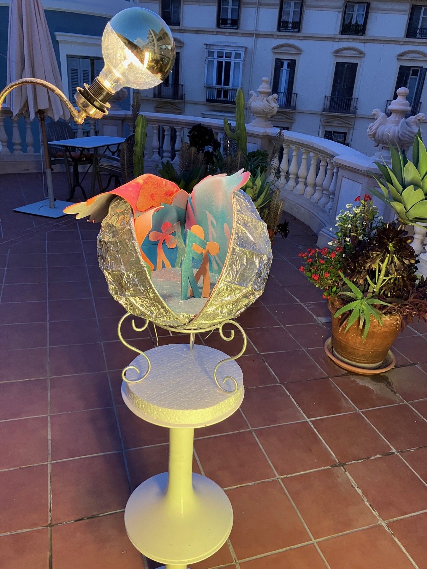 Dekorative Leuchte mit kugelförmigem Korpus auf einem weißen Hocker. Balkon mit Pflanzen und Gebäude im Hintergrund.