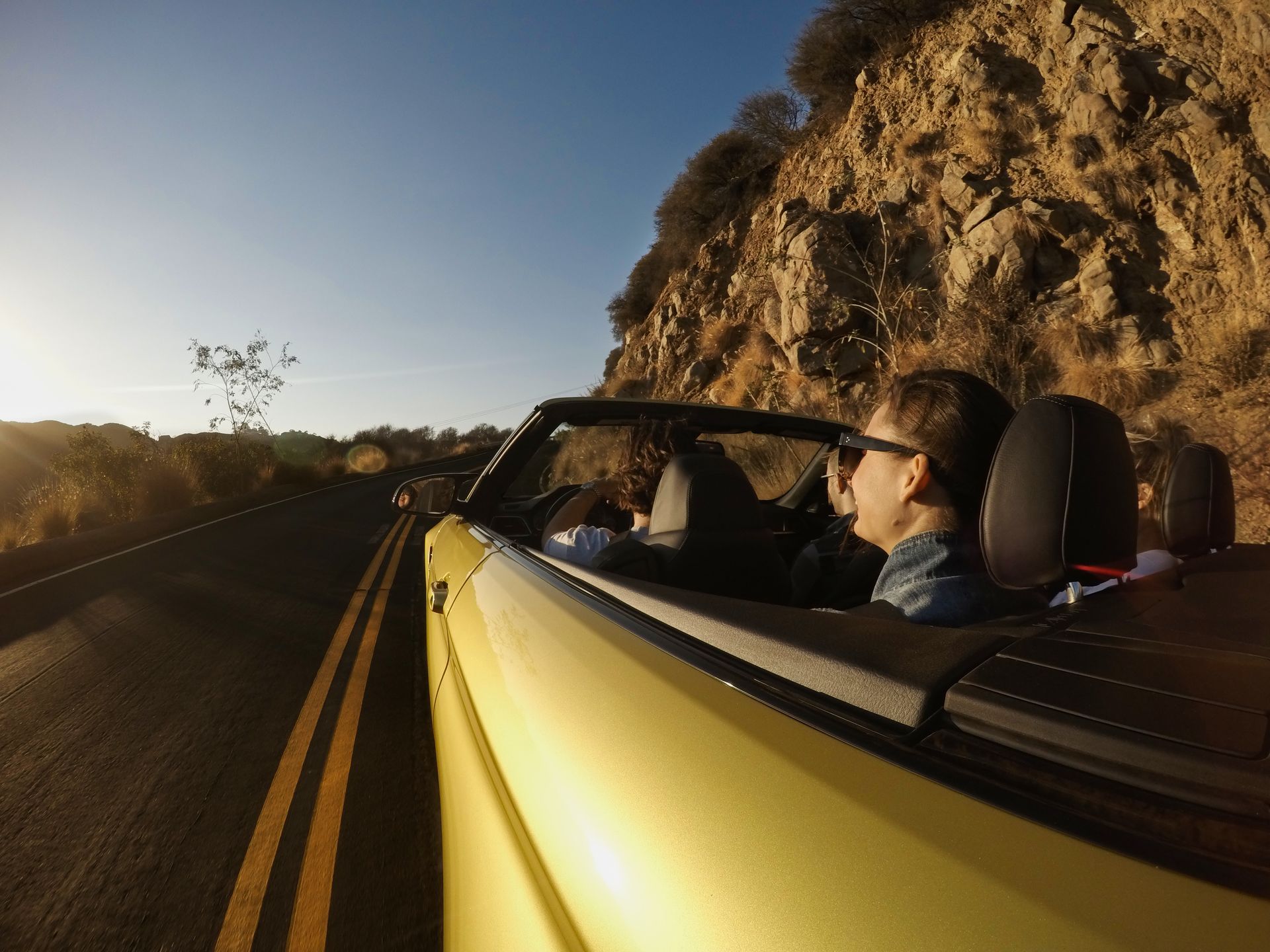 Two people in a yellow convertible driving on a winding road beside a rocky cliff at sunset