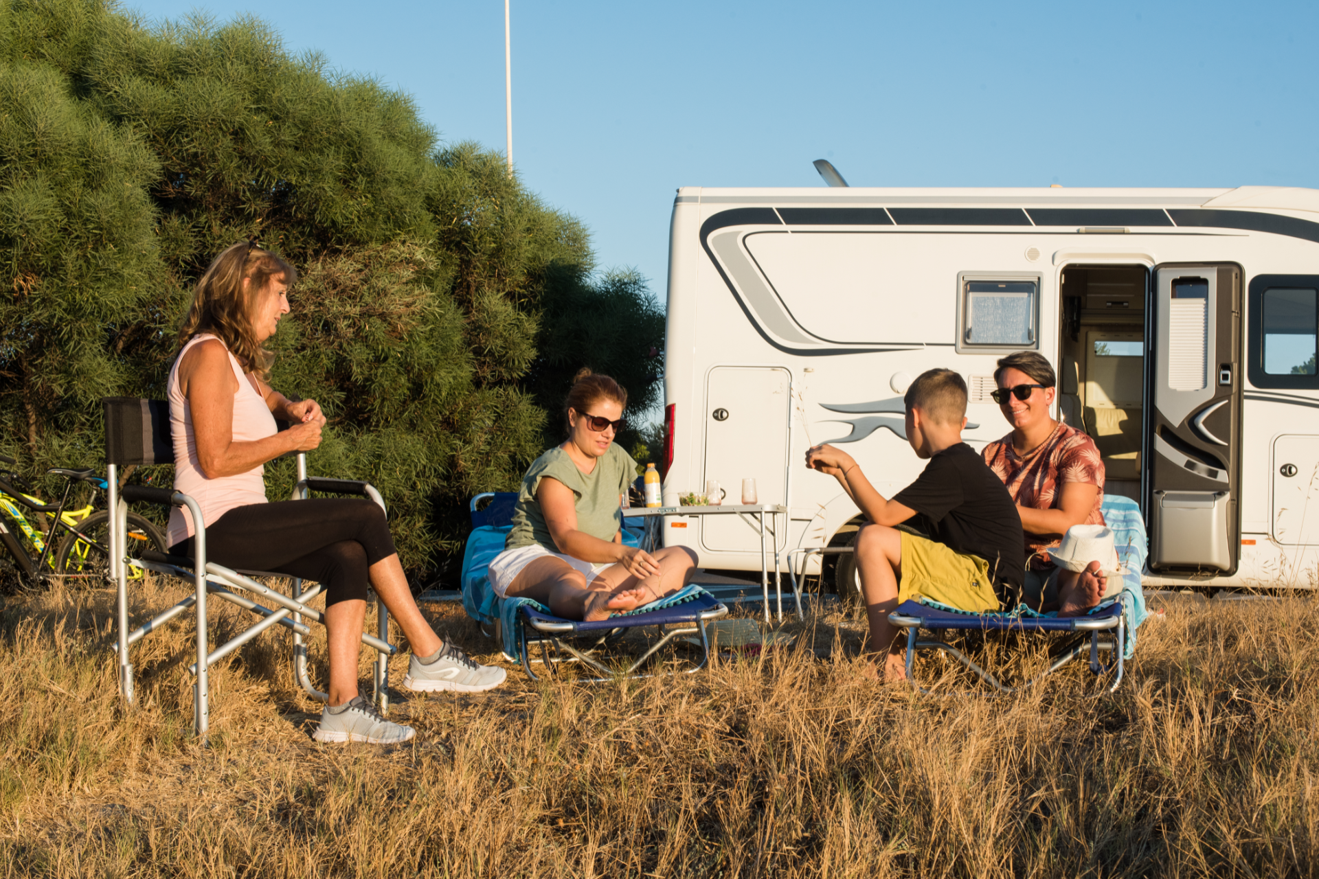 People relaxing outdoors beside a camper van, seated in foldable chairs on dry grass.