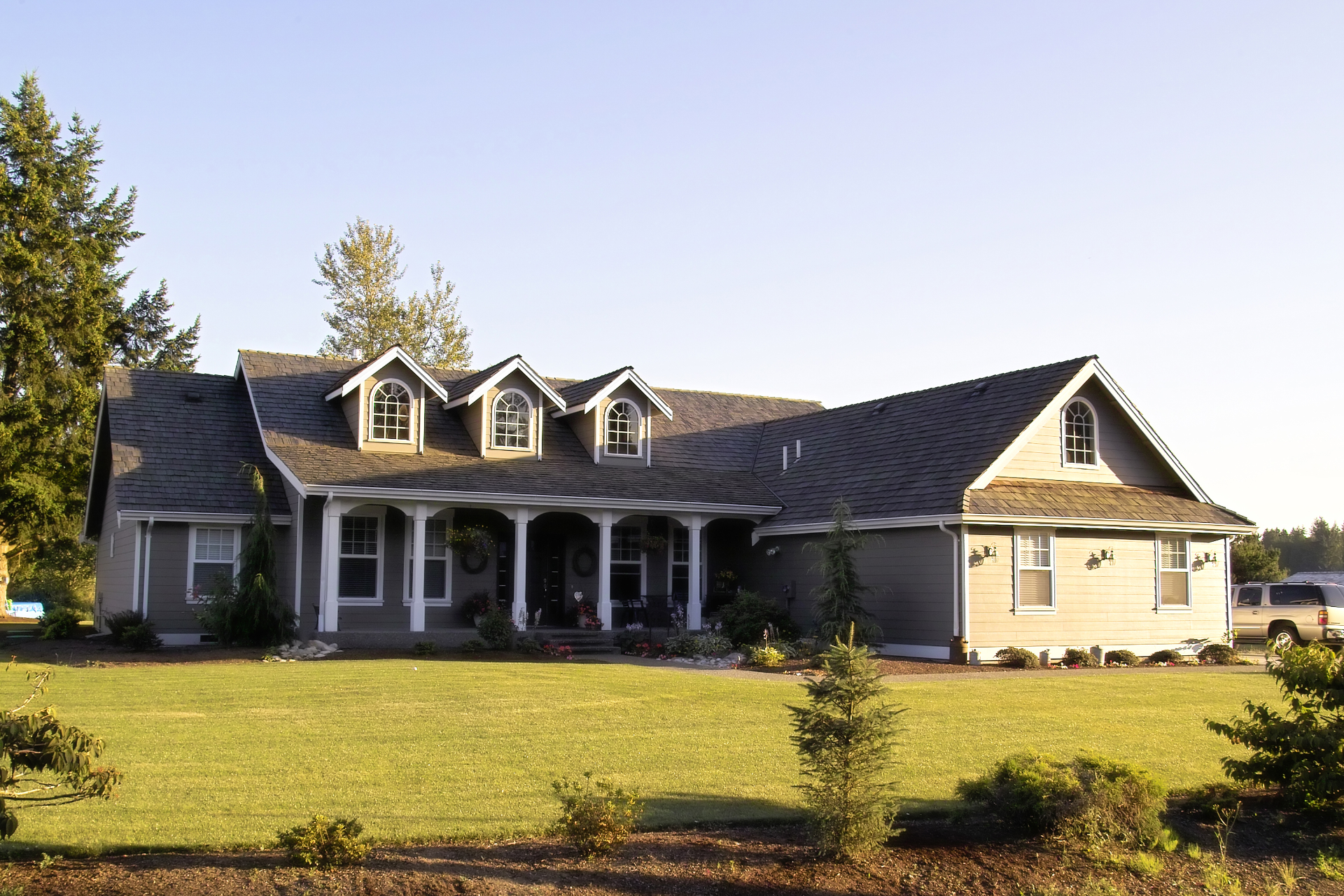 Large gray suburban house with a covered porch, dormer windows, and a wide lawn in daylight