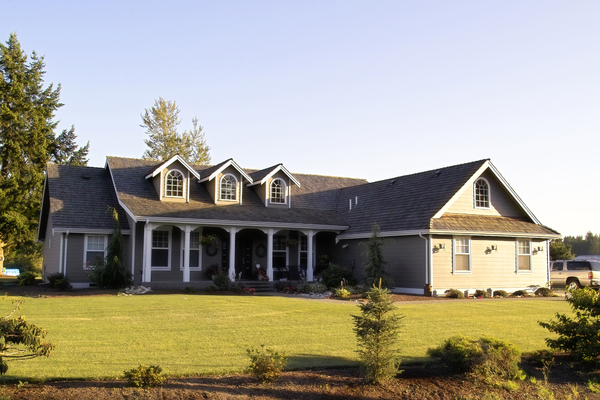 Large gray suburban house with a covered porch, dormer windows, and a wide lawn in daylight