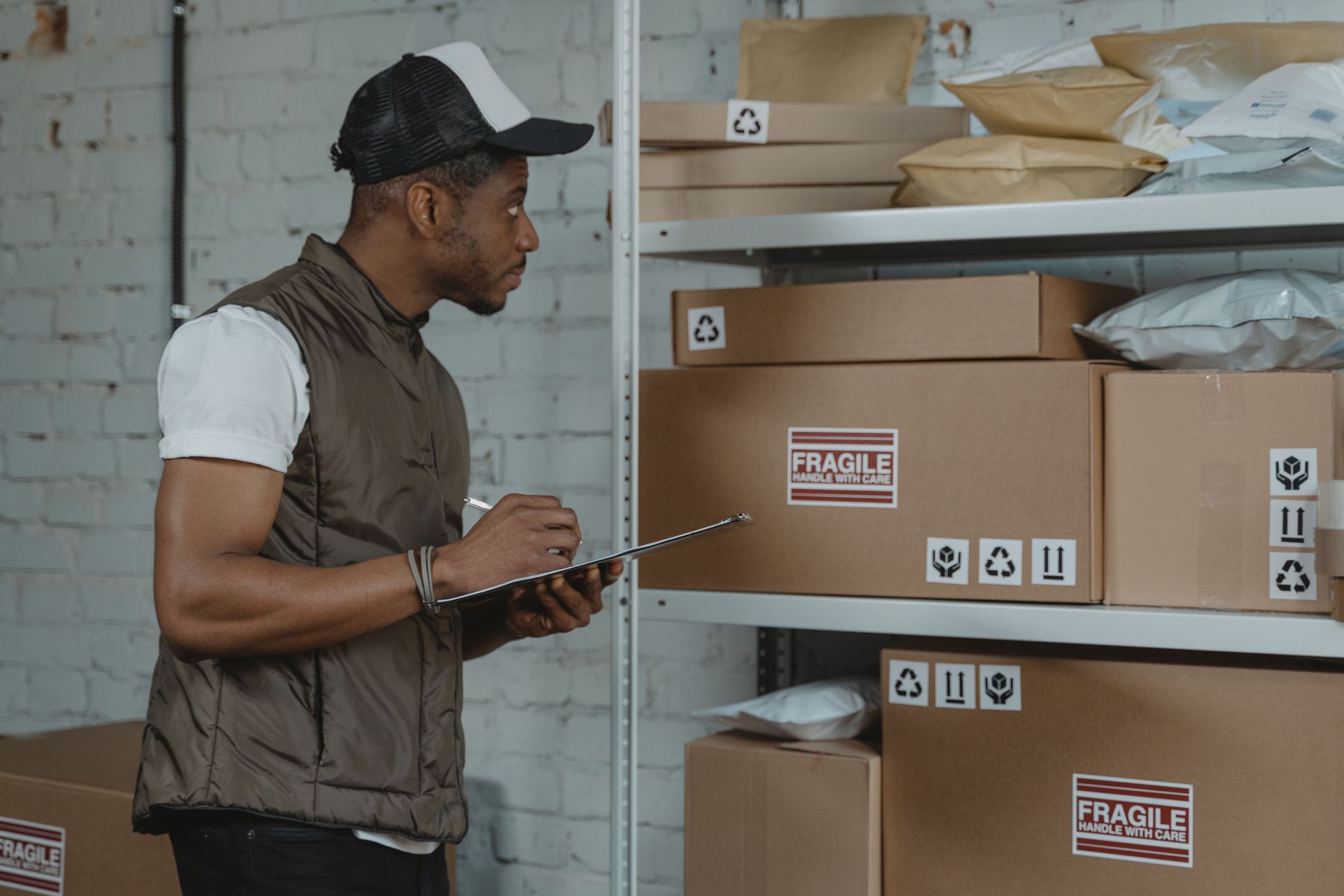 Man examining labeled cardboard boxes on warehouse shelves, holding a clipboard.