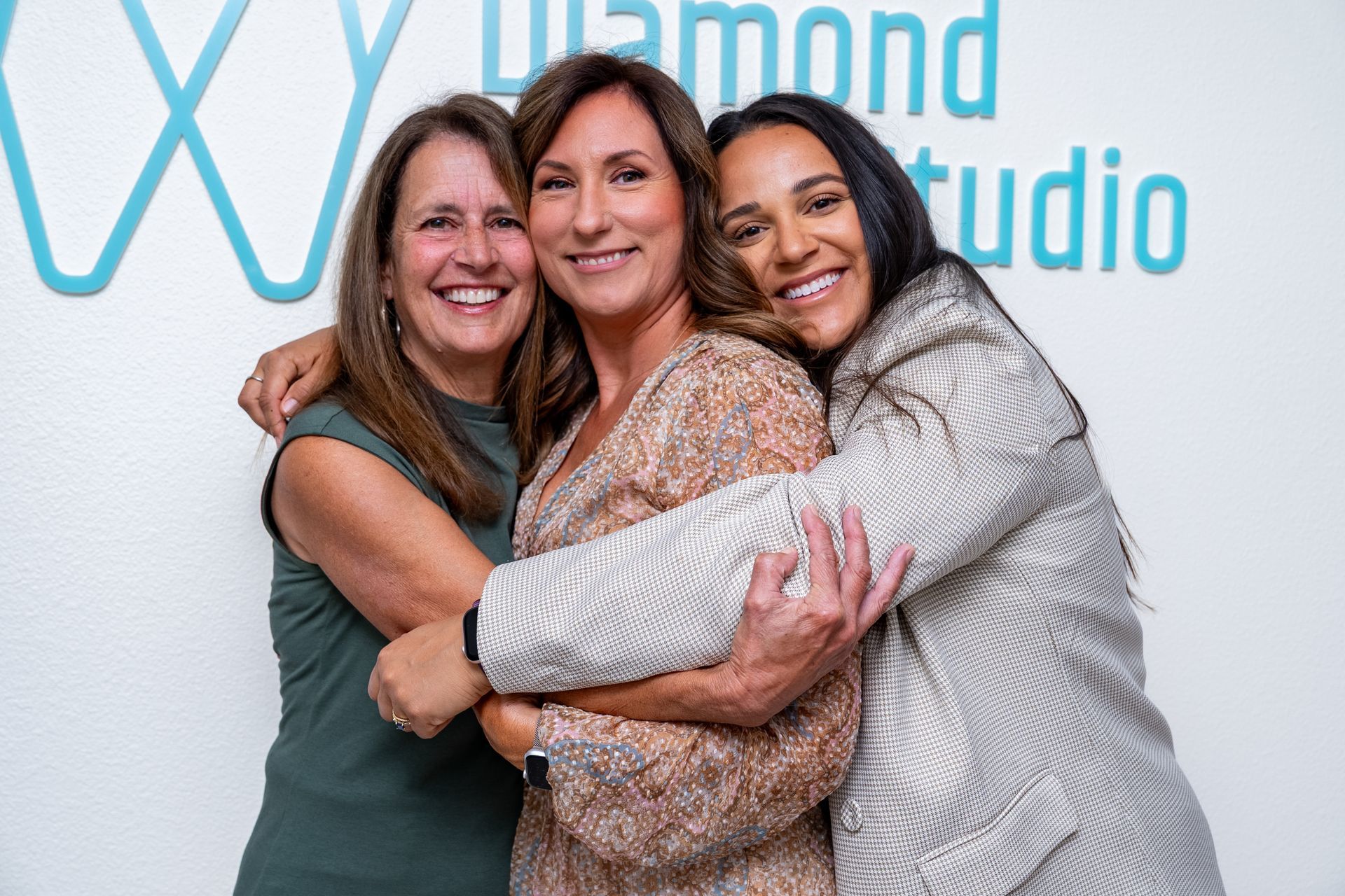 Three smiling women embracing in front of a white wall with 