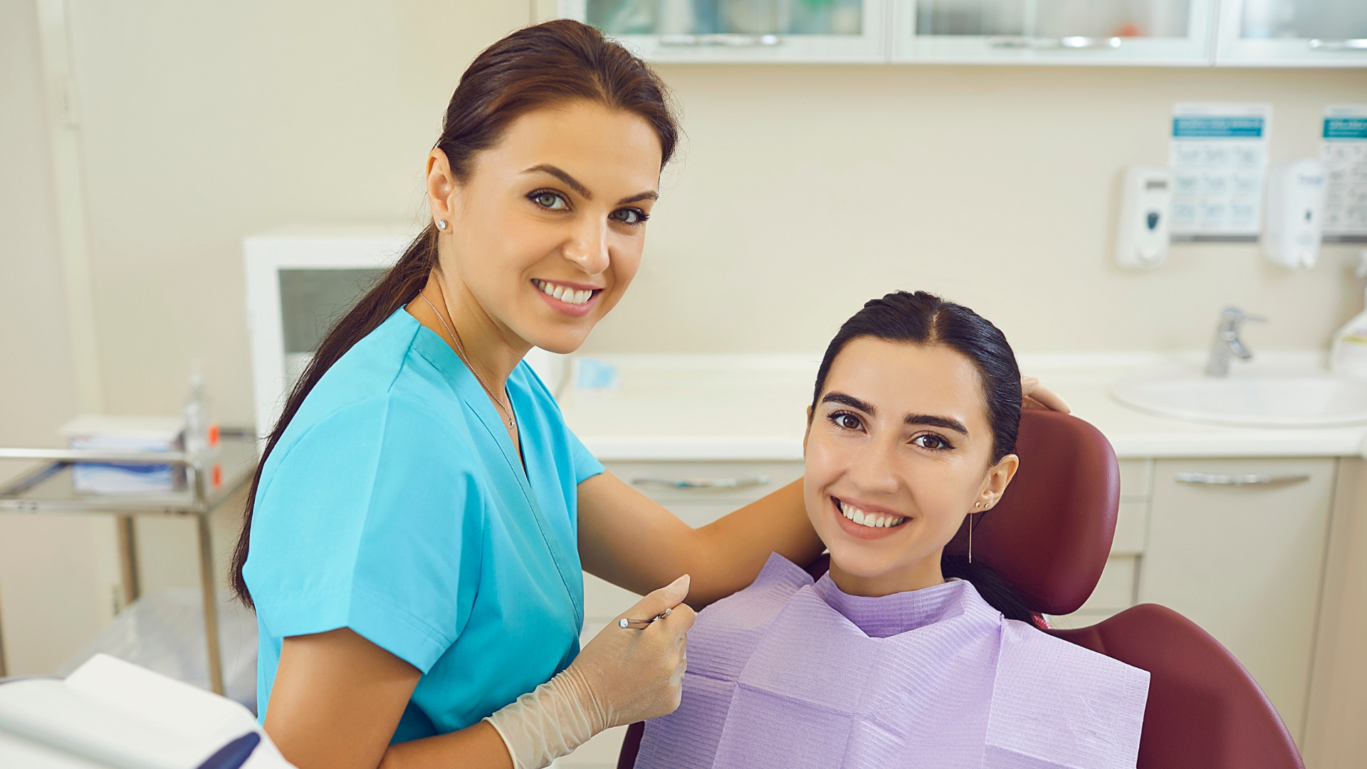 Dental assistant smiling beside a patient in a clinic, both in scrubs and a bib-covered chair