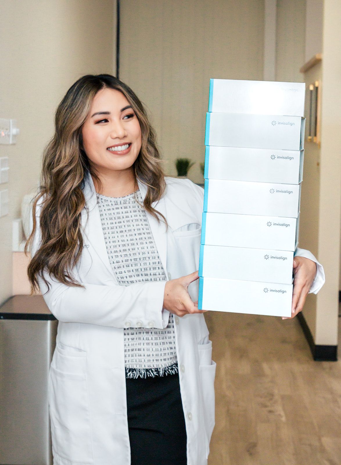 Woman in lab coat holding a stack of white boxes in a medical office, smiling.