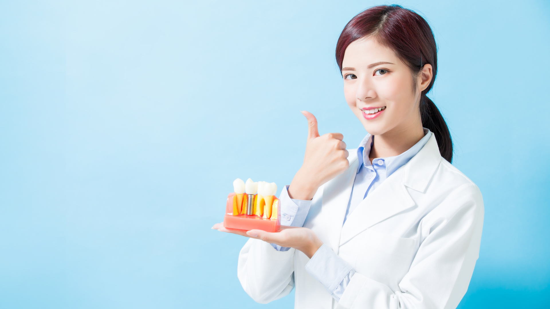 Smiling dentist in a white coat holds a tooth model, giving a thumbs-up. Blue background.