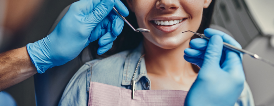 A dentist examining a patient's teeth with tools. Hands in blue gloves are in focus. The patient smiles.