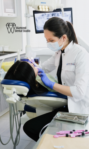 Dentist in white coat and mask examines a patient in dental chair, light blue gloves, dental tools on tray.