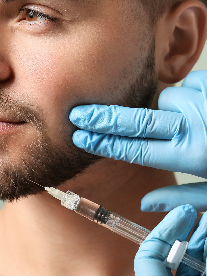 A man receiving a facial injection in a medical setting. Blue-gloved hands hold syringe to his cheek.