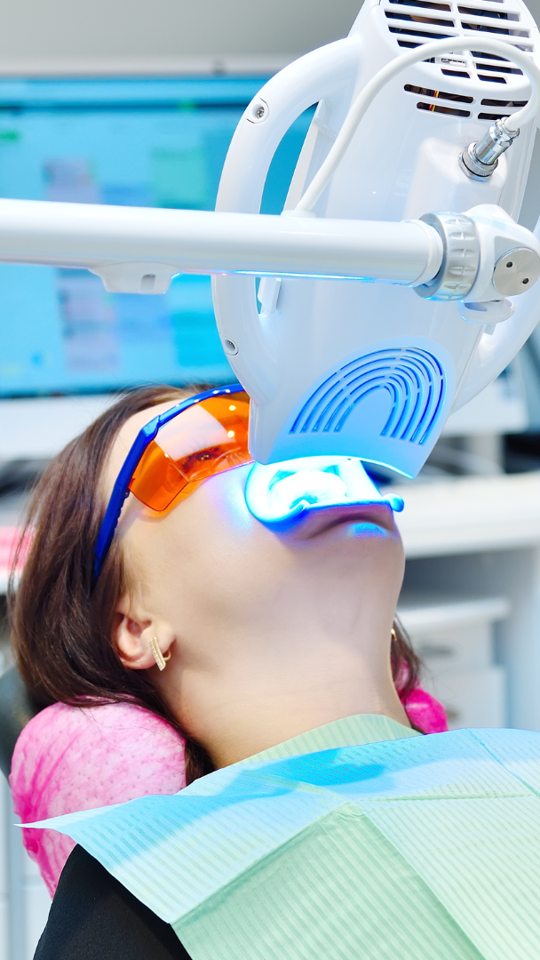 Woman undergoing teeth whitening with blue light, wearing protective orange glasses, in a dental office.